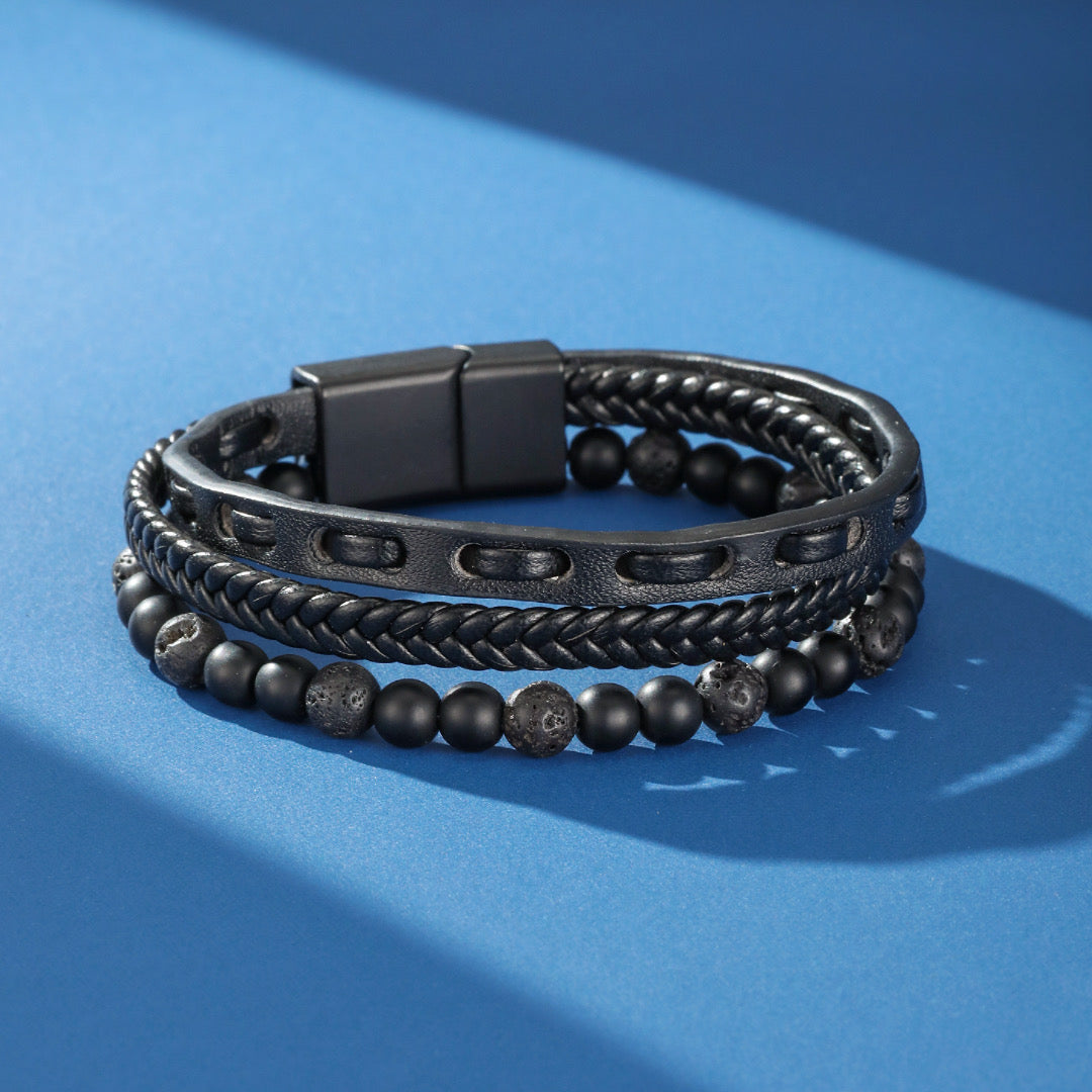 Bracelet multicouche pour homme en cuir tressé et perles de lave noire, doté d'un fermoir magnétique noir sur fond bleu avec des ombres.