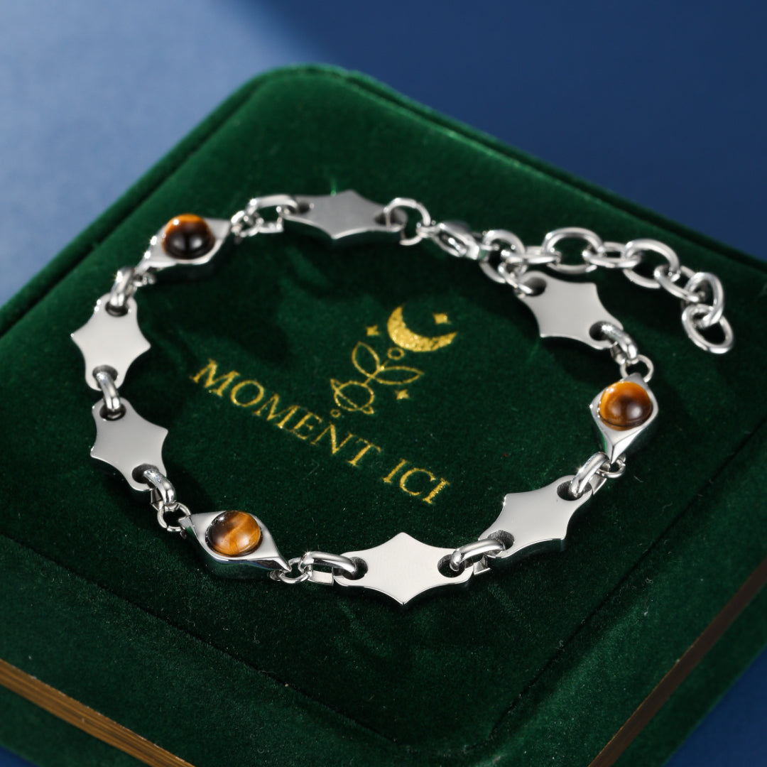 Bracelet pour homme en argent avec des pierres d'œil de tigre et des maillons argentés en forme de losange, sur un écrin en velours vert avec le logo de la marque.