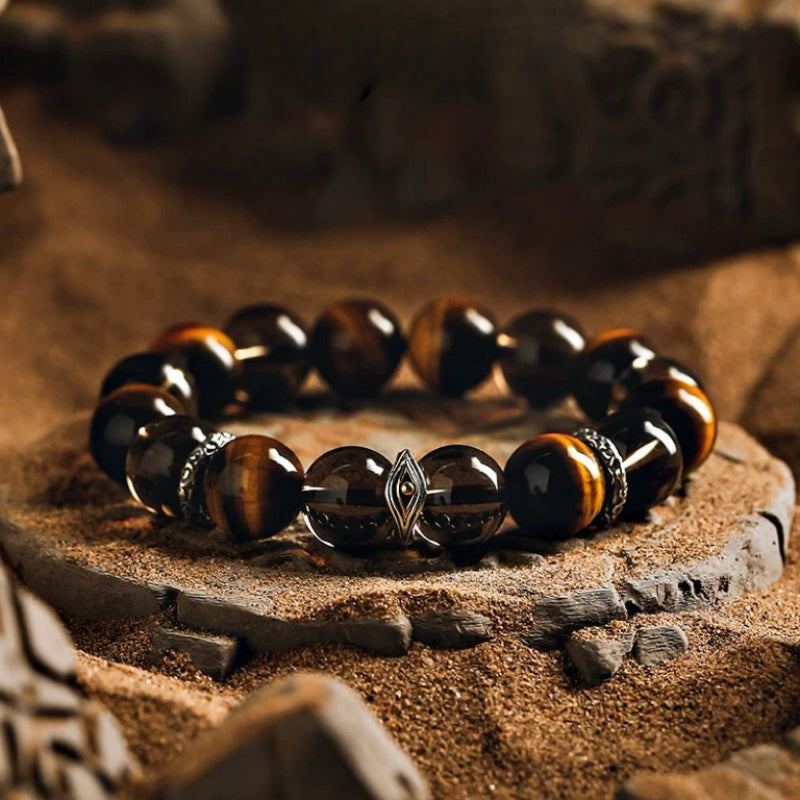 Bracelet pour homme avec perles œil de tigre, œil de protection et détails argentés, posé sur un lit de sable.