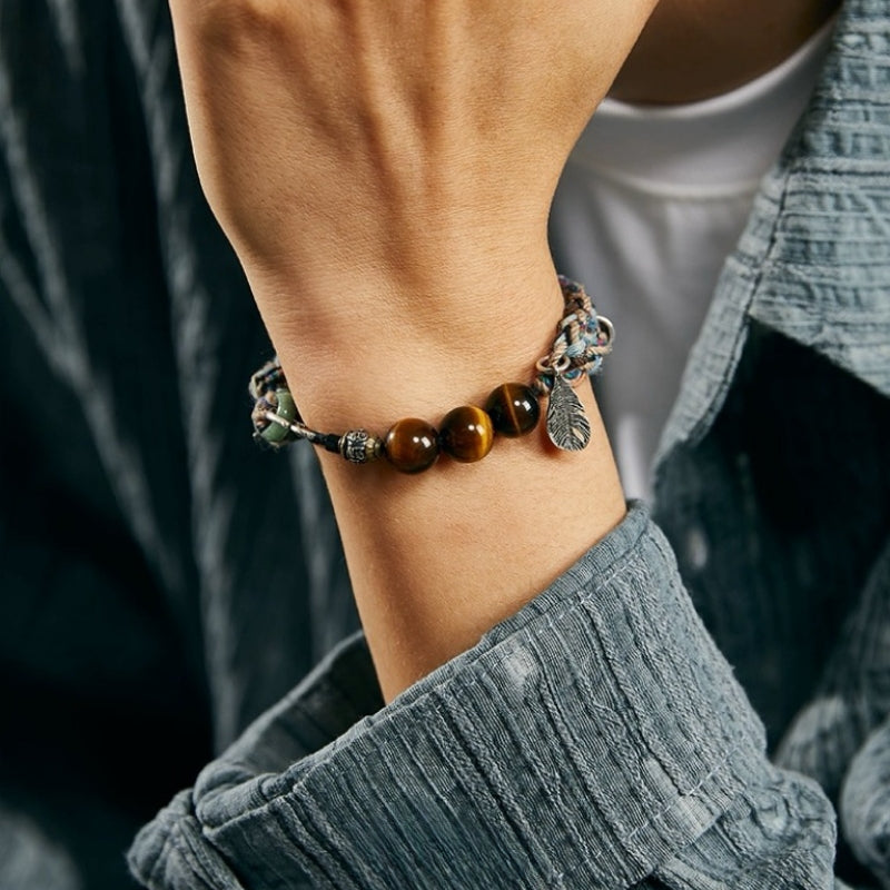 Bracelet en œil de tigre avec des perles marron et un pendentif plume en argent, porté au poignet d'une personne.