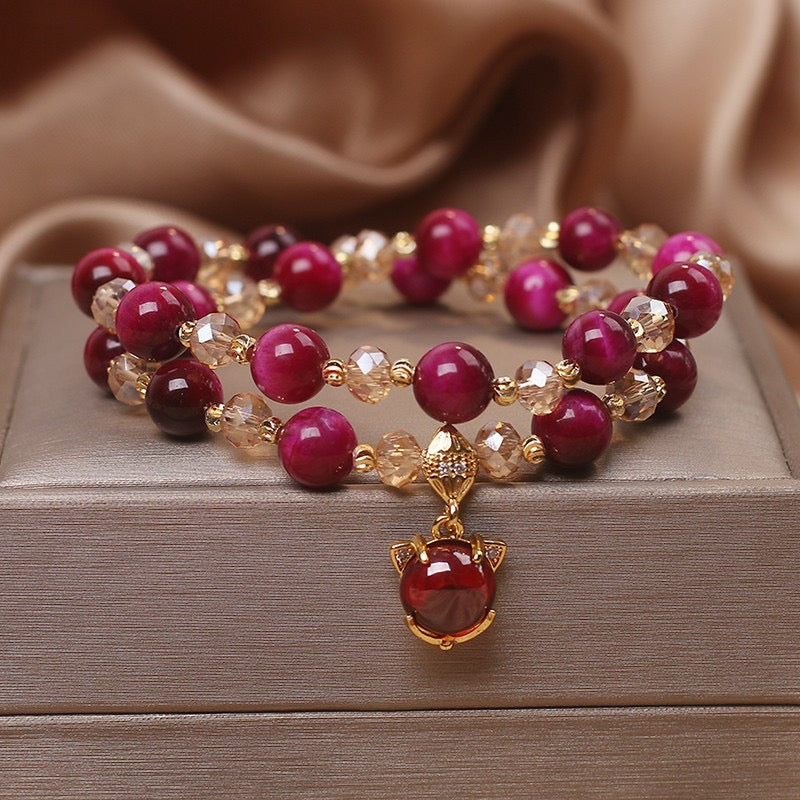 Bracelet double rang avec perles rondes de couleur bordeaux et perles facettées dorées, orné d'un pendentif en forme de tête de chat stylisée avec une pierre rouge centrale, monté sur une chaîne plaqué or.