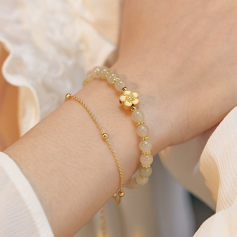 Bracelet fait de perles de jade pâle et d'une breloque fleur dorée, porté à l'avant-bras, avec un autre bracelet chaîne fine dorée.
