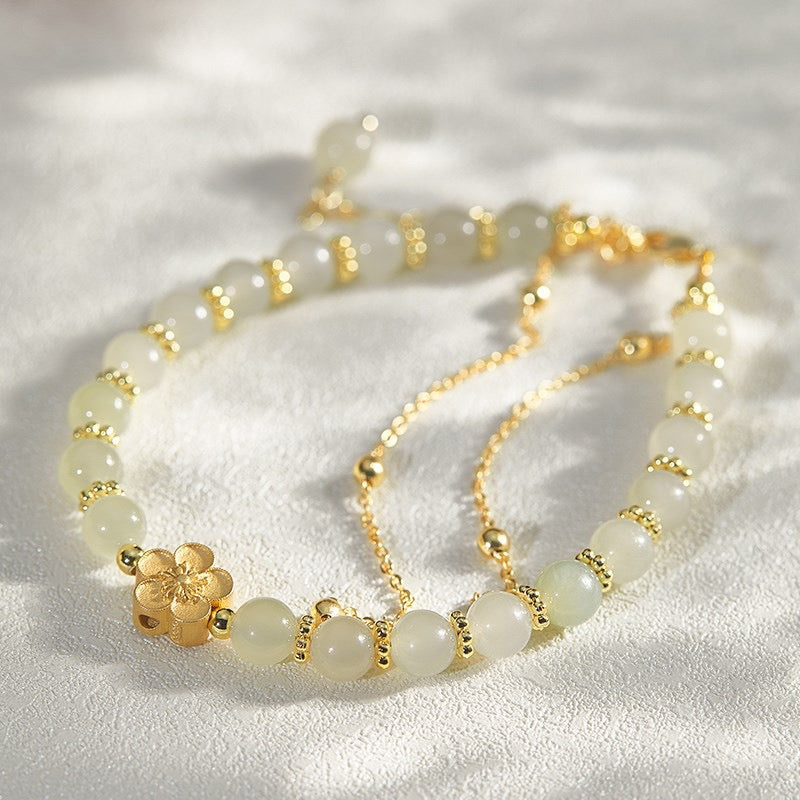 Bracelet en jade pâle avec des perles dorées et un pendentif fleur en or, sur un fond blanc texturé.