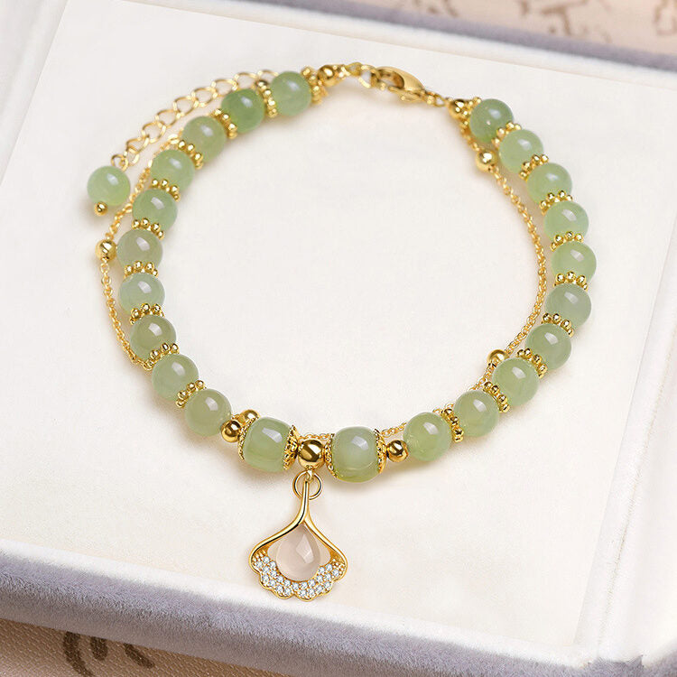 Bracelet double rang avec perles en jade vert clair, fermoir et chaîne dorés, et breloque feuille de ginkgo ornée de cristaux.