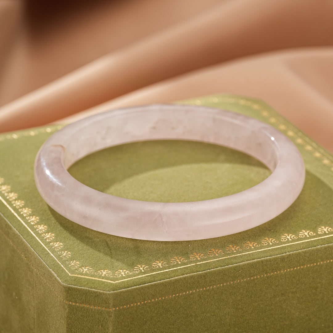 Bracelet jonc en quartz rose translucide, posé sur une boîte verte ornée de motifs dorés, avec un fond soyeux rose pâle.