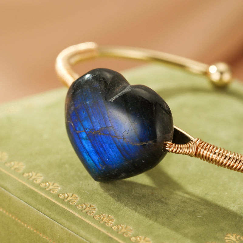 Bracelet jonc doré avec une pierre de labradorite en forme de cœur aux reflets bleus intenses et irisés, sur un fond vert