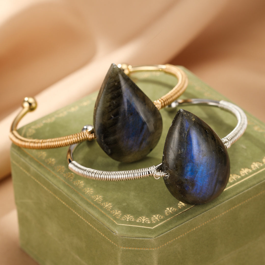 Deux bracelets jonc ornés de grosses gouttes de labradorite bleue et grise, montés sur des fils métalliques argentés et dorés, posés sur une boîte verte.