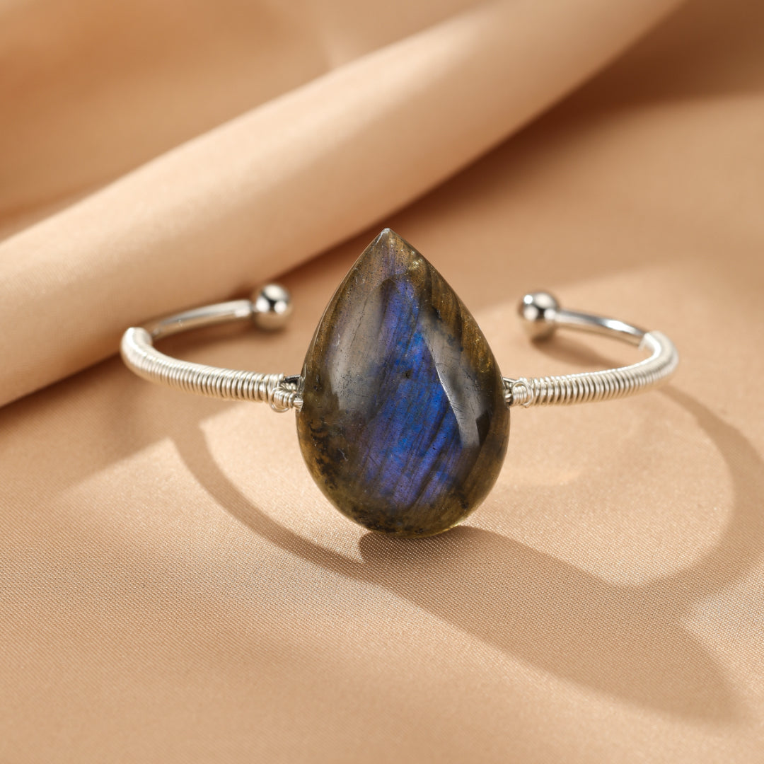 Bracelet jonc en argent avec une grosse pierre de labradorite en forme de gouttelette bleue et grise au centre, sur un fond beige.