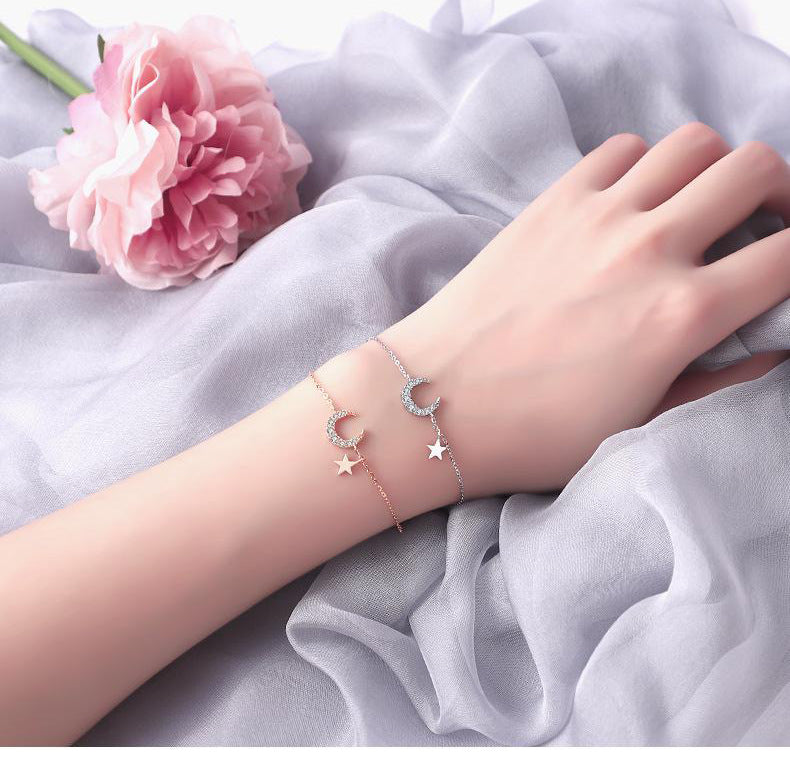 Deux bracelets fins en argent ornés de breloques lune et étoile scintillantes, portés sur un bras par-dessus un tissu gris doux, avec une fleur rose en arrière-plan.