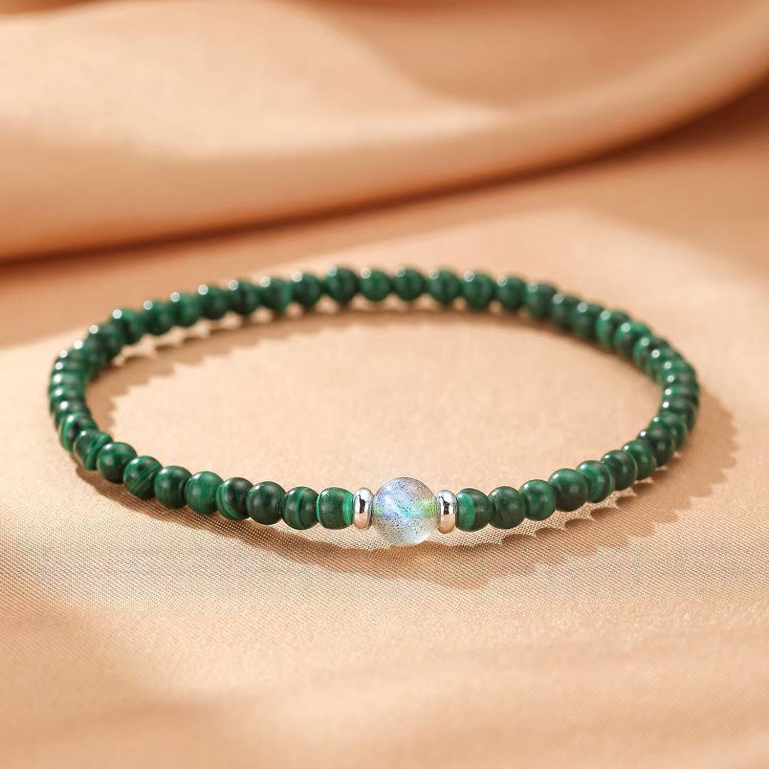 Bracelet en malachite avec une perle centrale irisée et des intercalaires argentés, sur un fond beige.