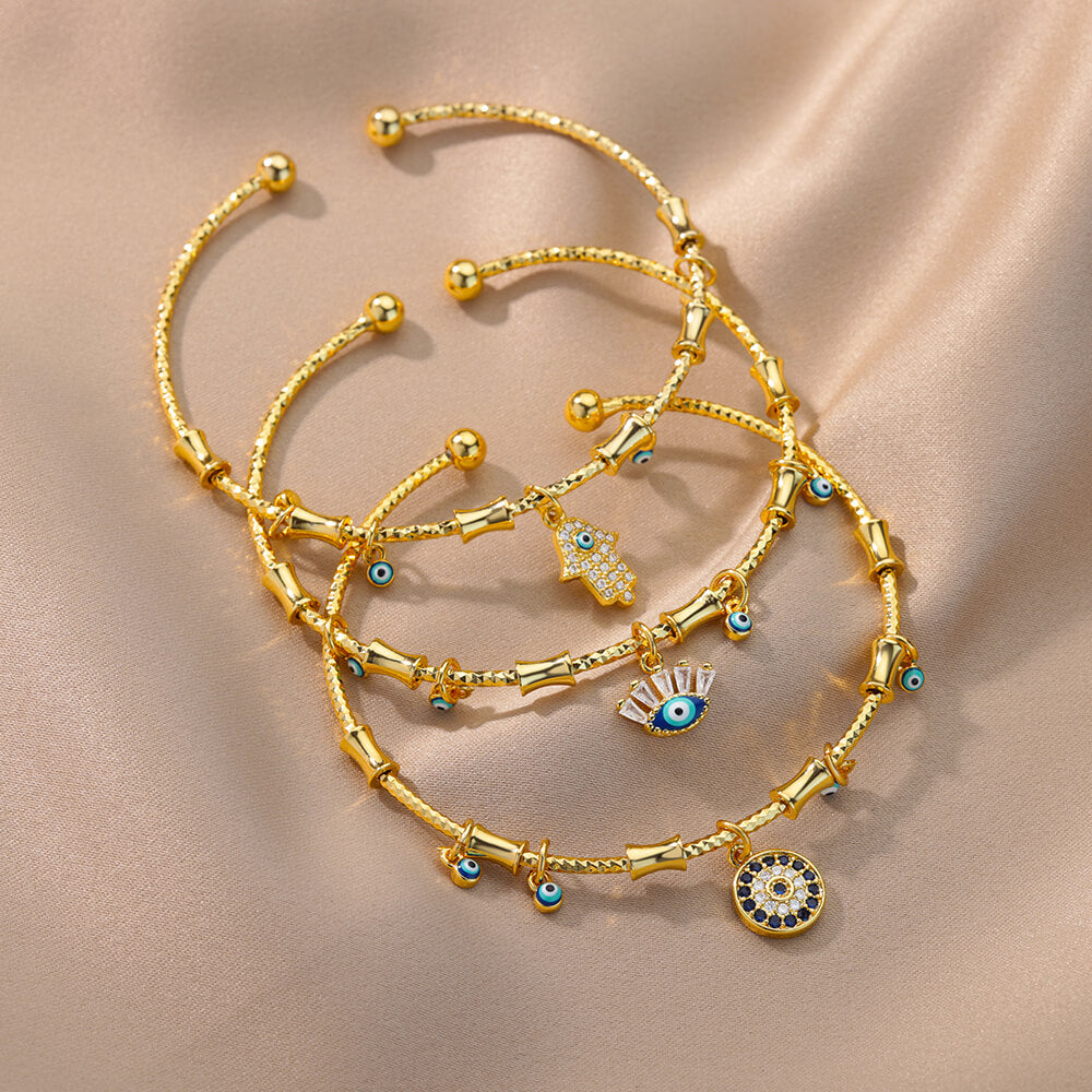 Trois bracelets plaqués or avec des breloques œil maléfique bleu, un œil de Fatima en zircon et une main de Fatima sertie de zircon, sur un fond satiné.