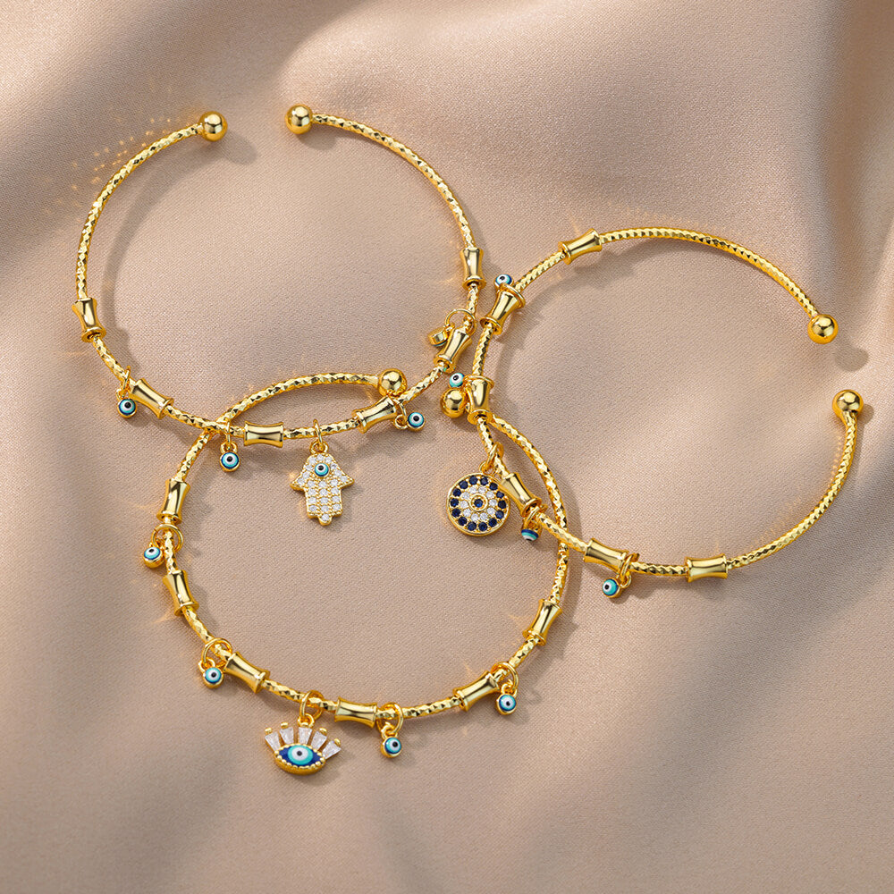 Trois bracelets dorés ornés de motifs mauvais œil, de mains de Fatma et de zircon bleu, sur fond beige