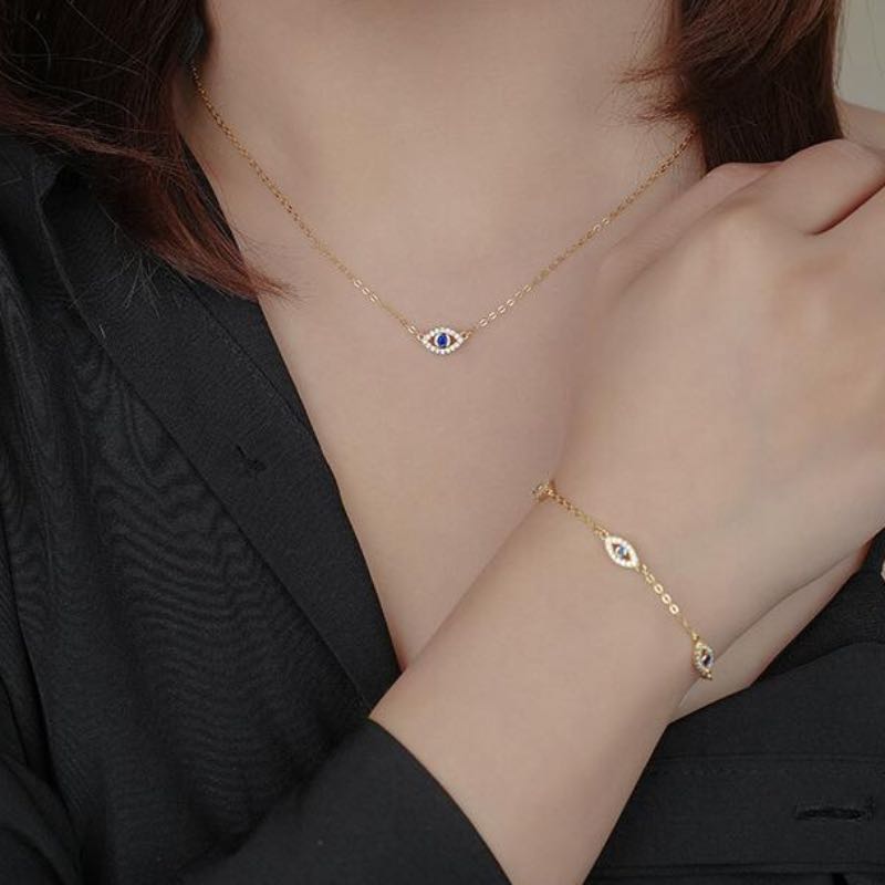 Gros plan d'un bracelet et d'un collier assortis en plaqué or, ornés de petits pendentifs œil bleu sertis de zircons cubiques blancs, portés par une femme sur fond noir.