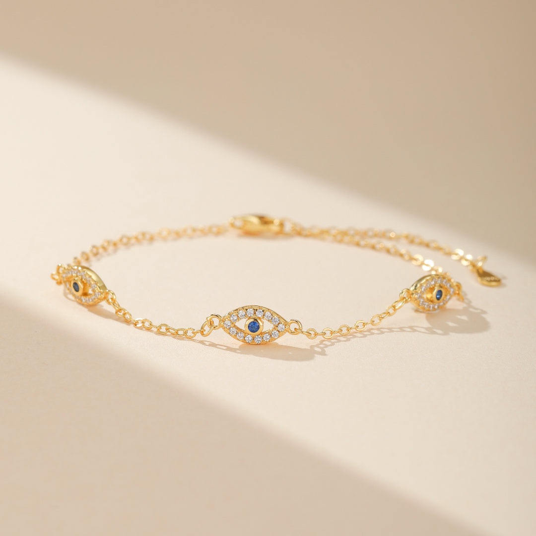 Bracelet en argent plaqué or orné de plusieurs breloques œil bleu serties de zirconias, sur fond beige clair avec des ombres douces.