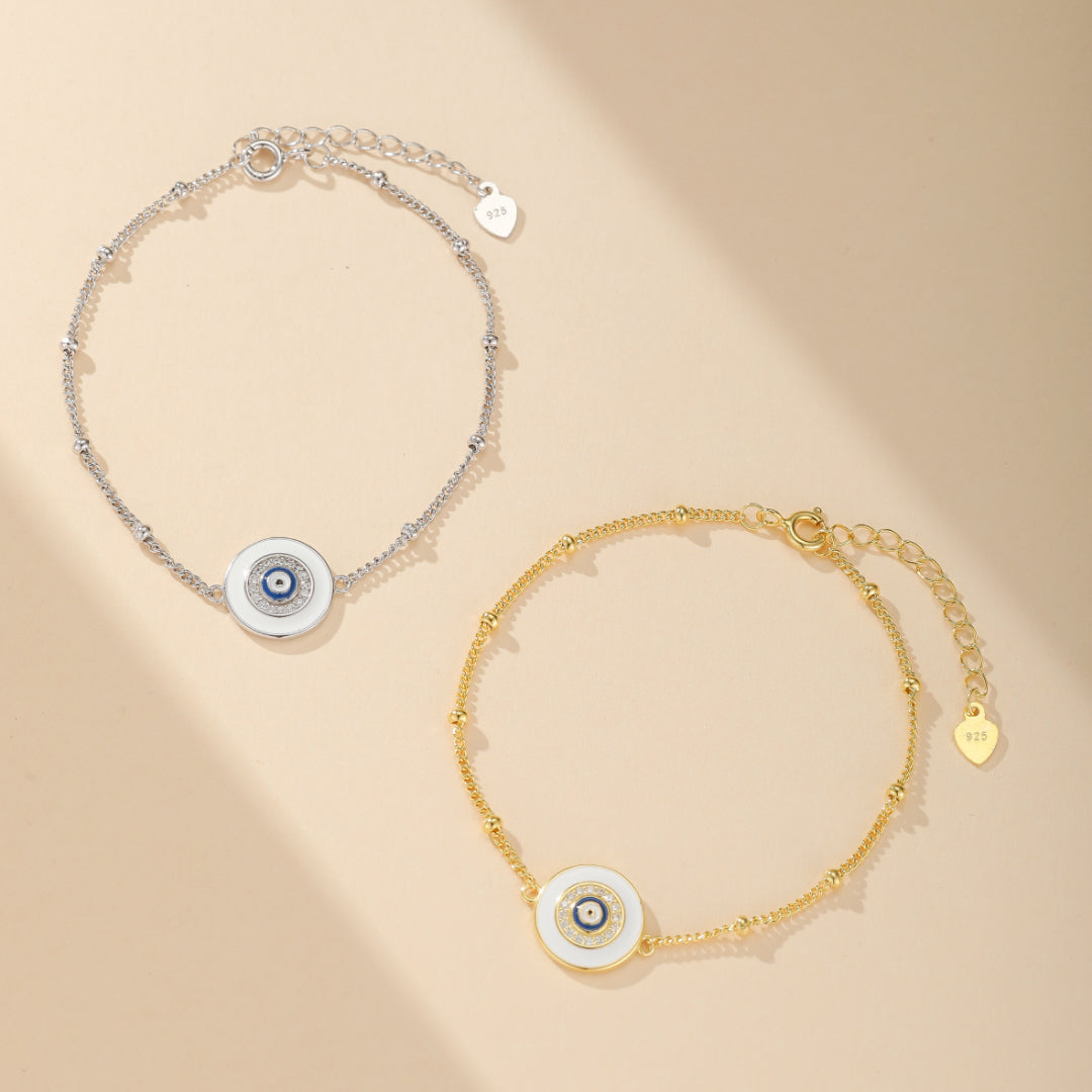 Deux bracelets avec un pendentif œil bleu et blanc, l'un en argent 925 et l'autre en plaqué or, sur fond beige.