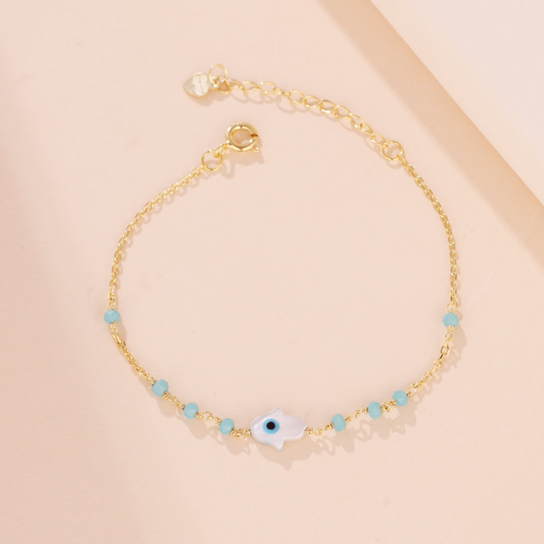 Bracelet en plaqué or avec une main de Fatma blanche ornée d'un œil bleu et de petites perles turquoise, symbolisant la protection céleste.