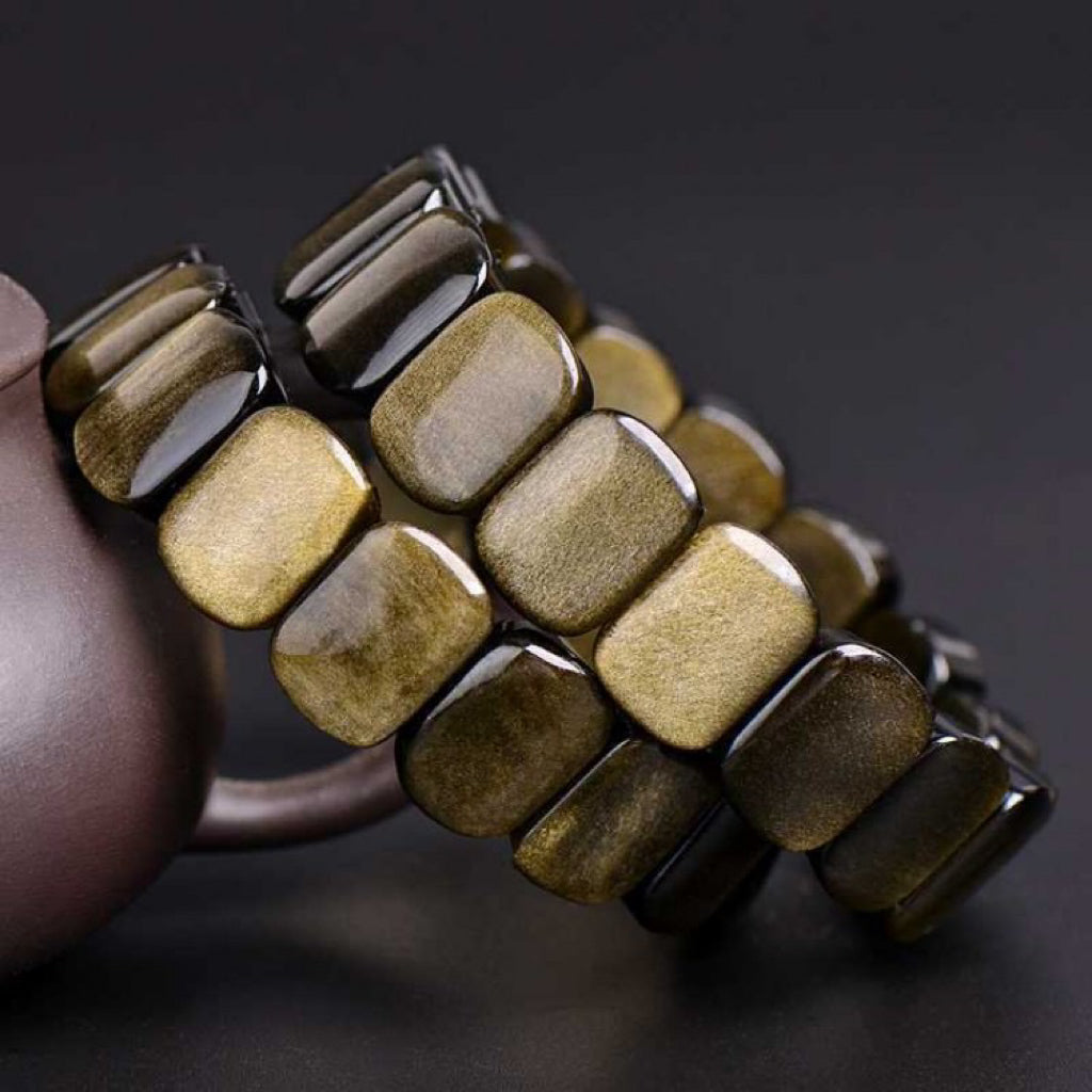 Bracelet fait de perles rectangulaires en obsidienne dorée, montées sur un élastique, sur un fond sombre.