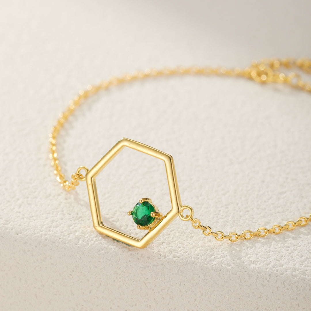 Gros plan sur un bracelet en plaqué or avec un pendentif en forme d'hexagone ajouré, orné d'une petite pierre verte ronde au centre. La chaîne est délicate et dorée.