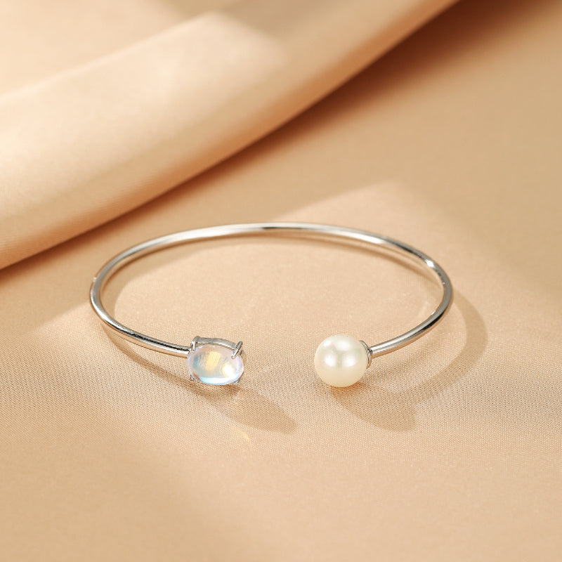 Bracelet jonc ouvert en argent 925, orné d'une pierre de lune lumineuse et d'une perle blanche, présentation sur fond satiné couleur pêche.