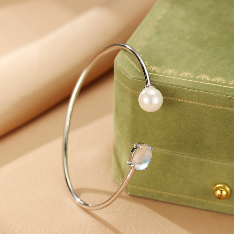Bracelet jonc ouvert en argent avec une perle blanche et une pierre de lune ovale bleutée, posé sur un écrin vert.
