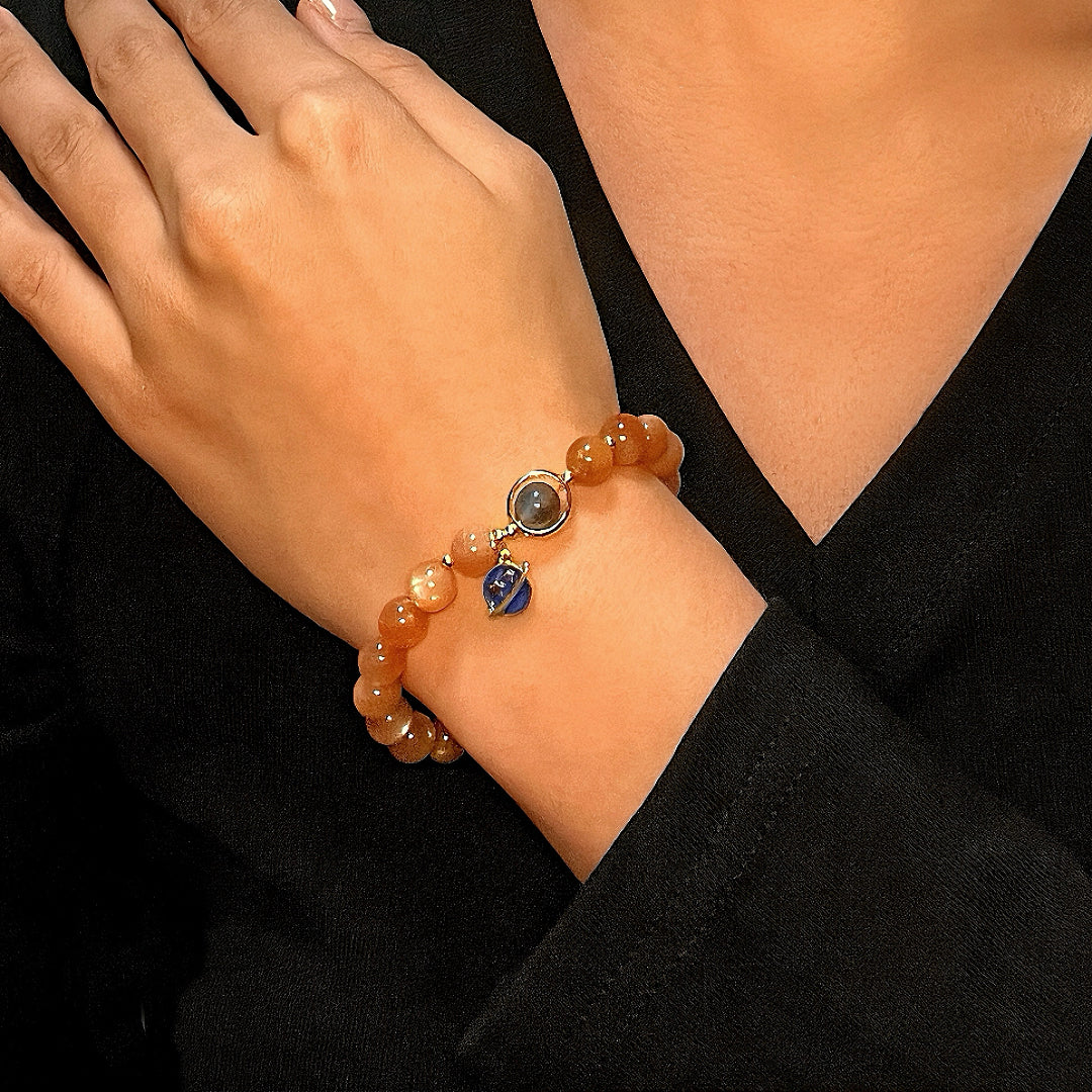 Gros plan d'un bracelet en pierre de soleil orange et en pierre de lune avec un pendentif bleu en forme de planète porté au poignet d'une femme.