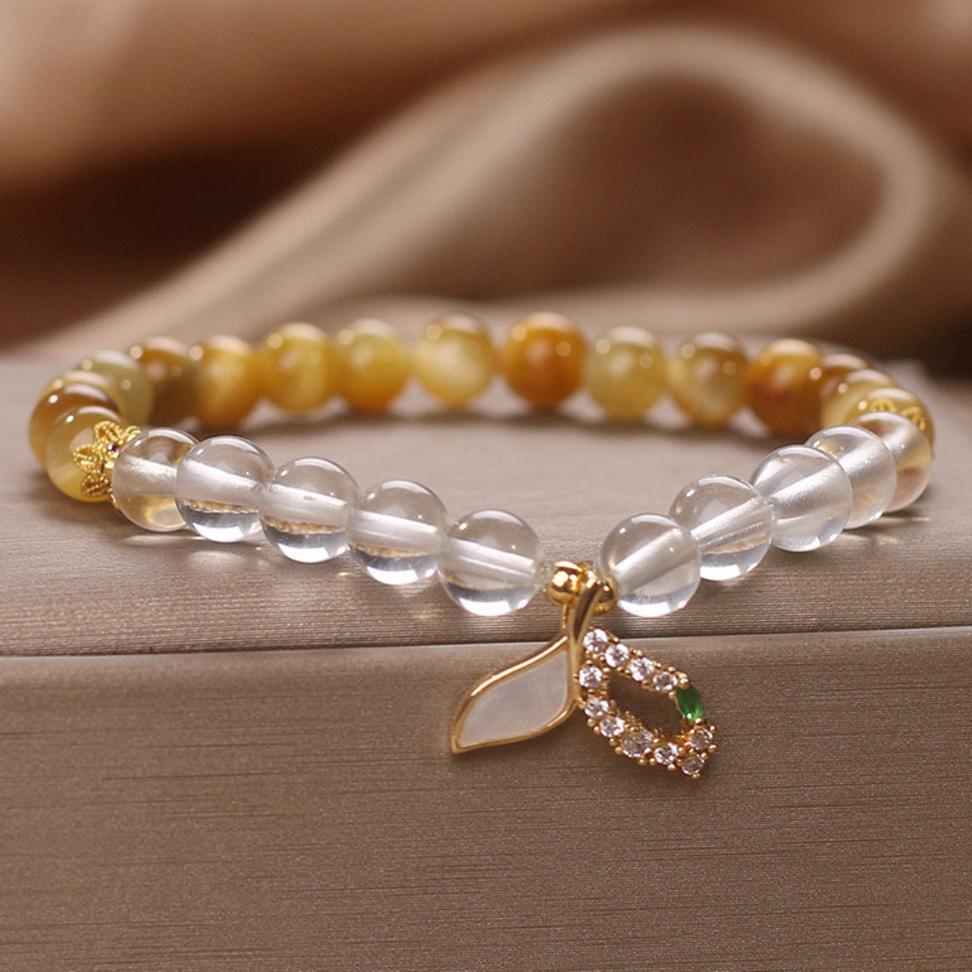 Bracelet élastique combinant des perles rondes en œil de tigre jaune et des perles transparentes en quartz clair, orné d'un pendentif feuille nacrée entouré de cristaux et d'une petite pierre verte.