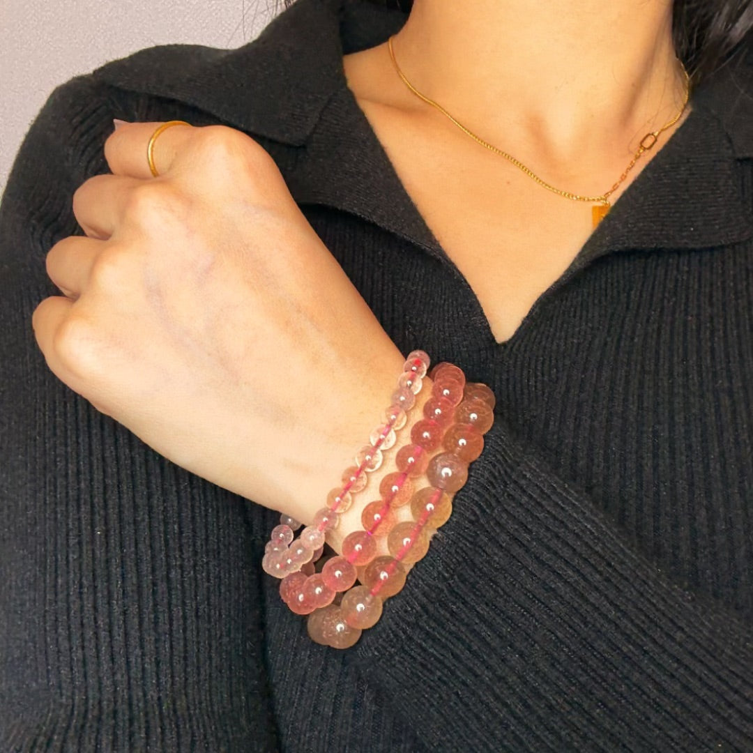 Trois bracelets en quartz fraise aux teintes rosées et translucides, portés à la main d'une femme sur un pull noir.