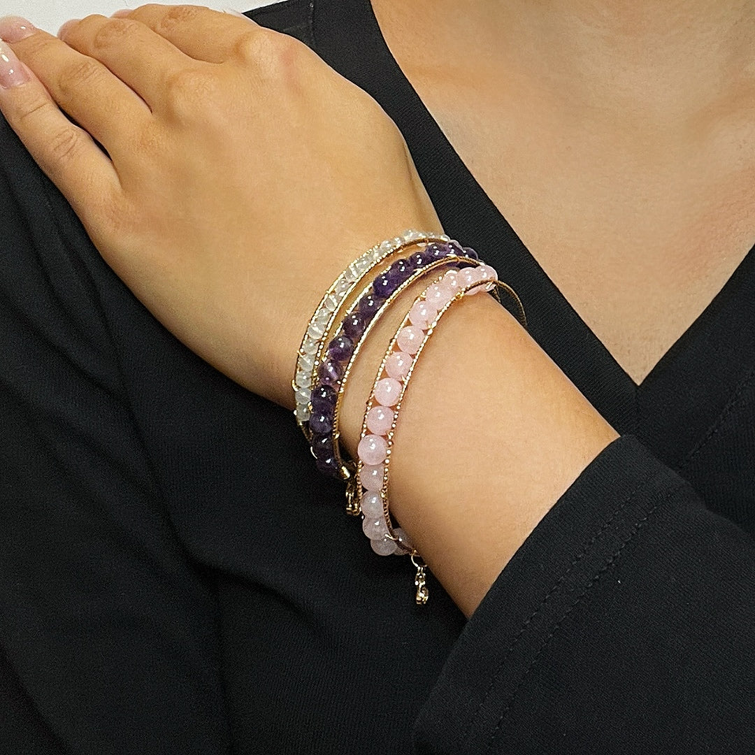 Trois bracelets empilables montés sur une monture dorée, ornés de perles de quartz rose, d'améthyste et d'aigue-marine, portés sur le poignet d'une femme.