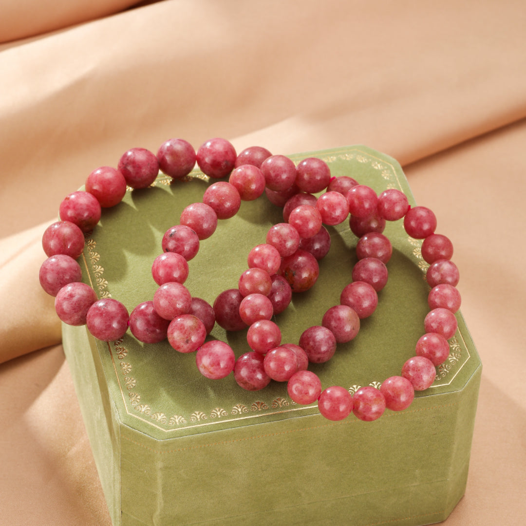 Deux bracelets en perles de rhodonite rose pâle disposés en cercle sur une boîte verte ornée d'une bordure dorée.