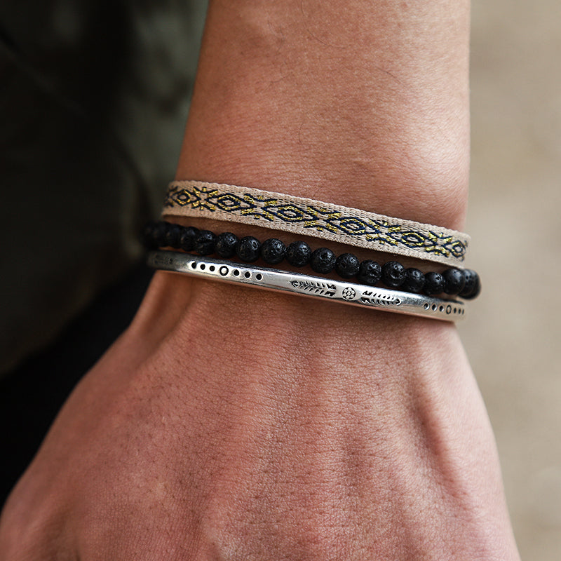 Ensemble de trois bracelets pour homme : un bracelet en tissu ethnique beige et noir, un bracelet en perles de lave noires et un bracelet manchette en argent gravé.