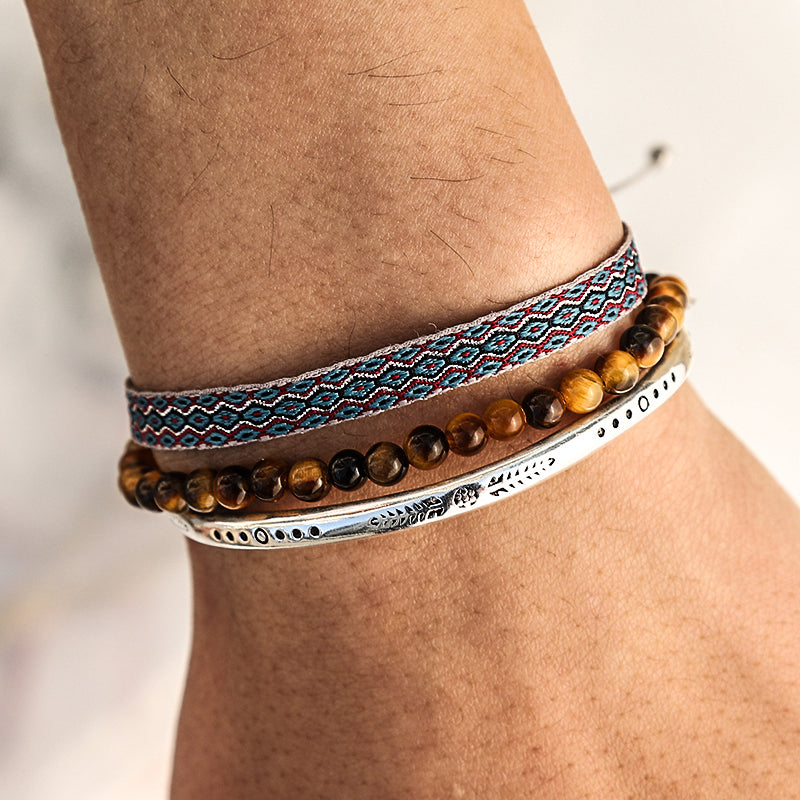 Ensemble de trois bracelets ethniques porté sur un avant-bras : un bracelet en perles d'œil de tigre, un bracelet jonc argenté gravé et un bracelet tressé bleu et rouge.