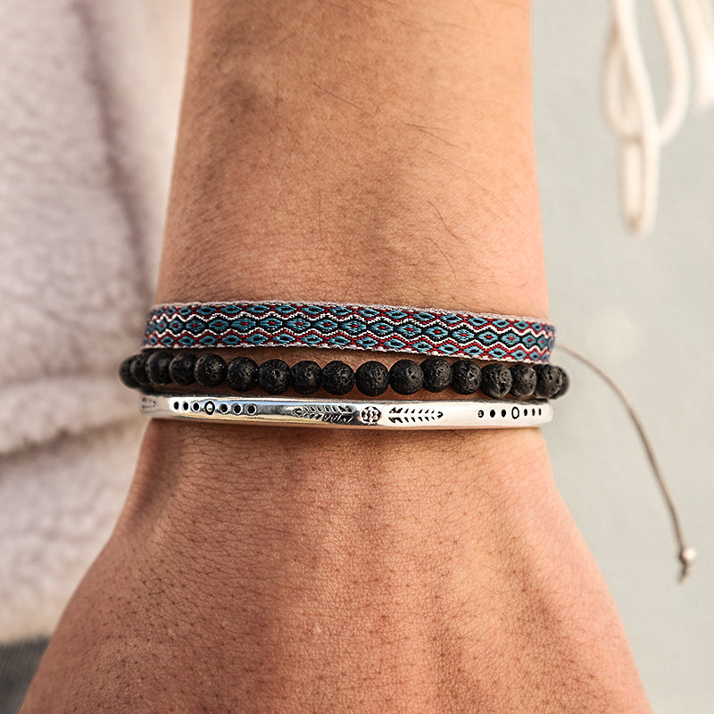 Ensemble de trois bracelets ethniques pour homme portés sur un poignet : un bracelet en tissu bleu et rouge, un bracelet de perles de lave noire et un bracelet jonc argenté gravé de motifs.
