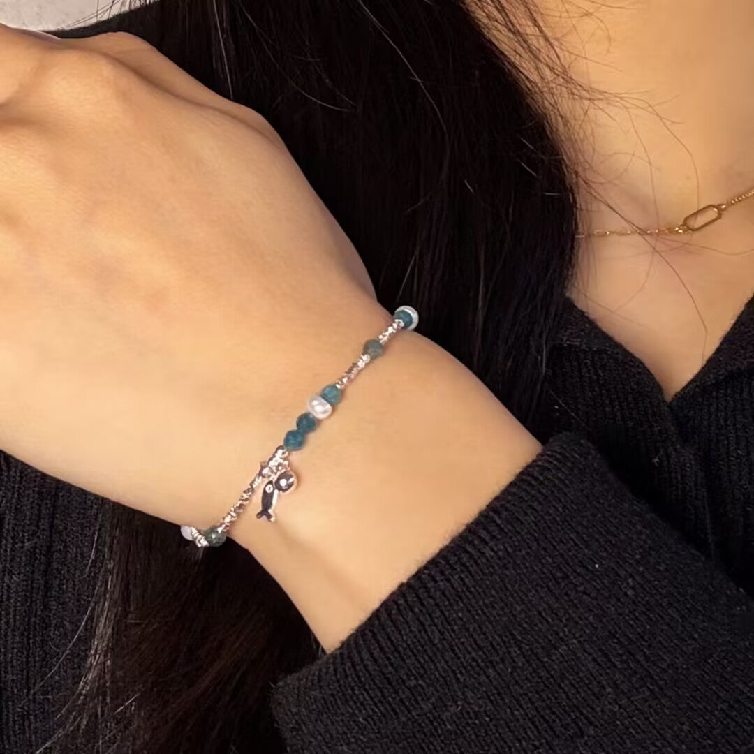 Bracelet fin en argent 925 avec des perles bleues et blanches et un charmant pendentif poisson d'azur, porté au poignet d'une femme.