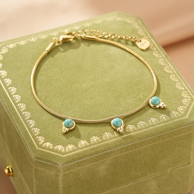 Bracelet en or jaune avec trois cabochons de turquoise disposés le long d'une chaîne serpent délicate, posé sur une boîte à bijoux verte.