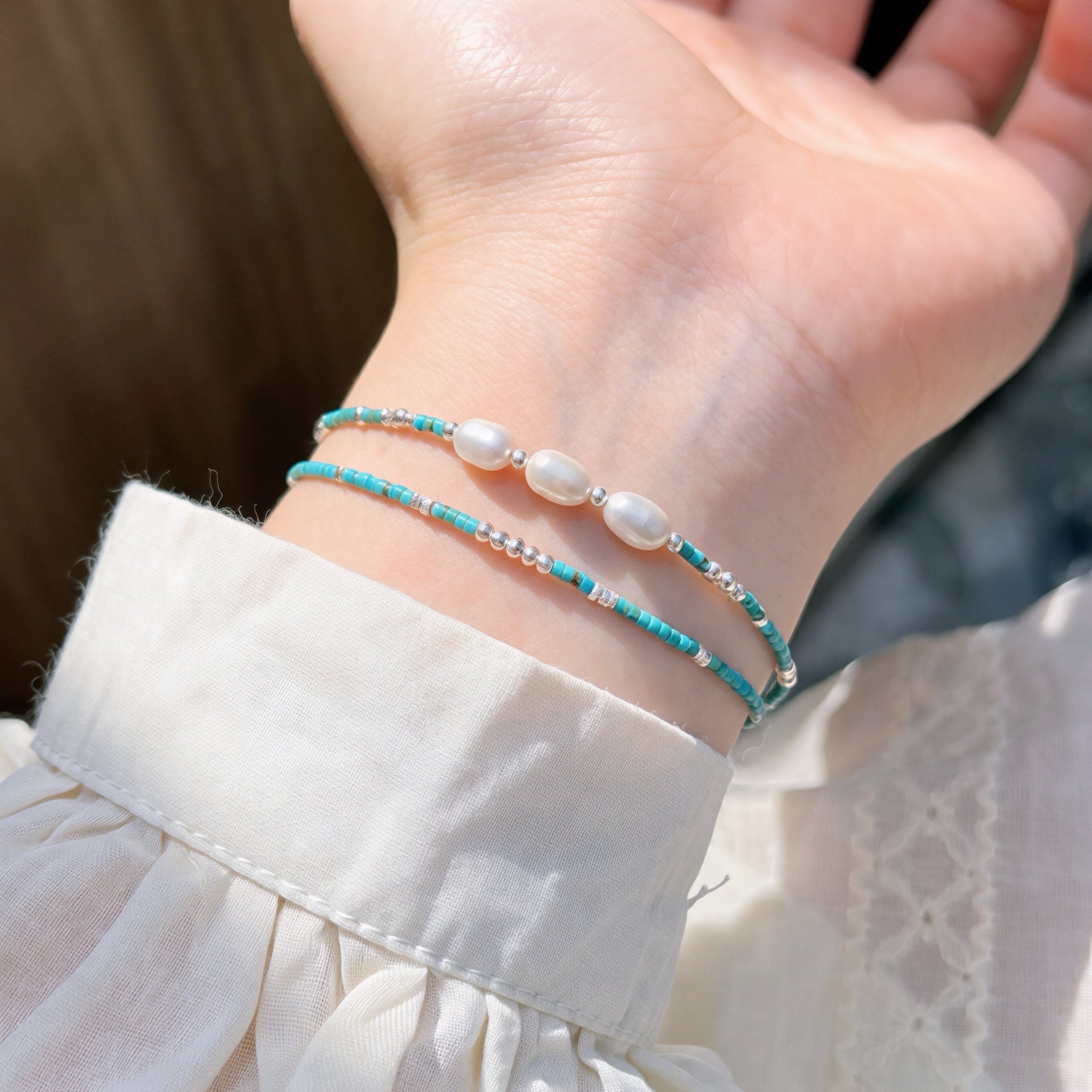 Deux bracelets fins en argent "Talisman du Vent" décorés de perles d'eau douce blanches et de petites pierres bleues turquoises, portés sur un poignet habillé d'une chemise blanche boutonnée et plissée.