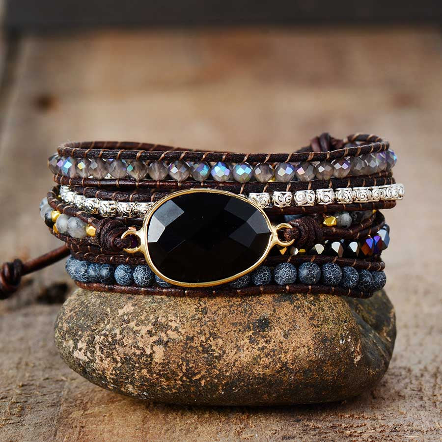 Bracelet enveloppant 'Force' en onyx avec des perles grises et bleues, monté sur du cuir marron et posé sur une pierre.