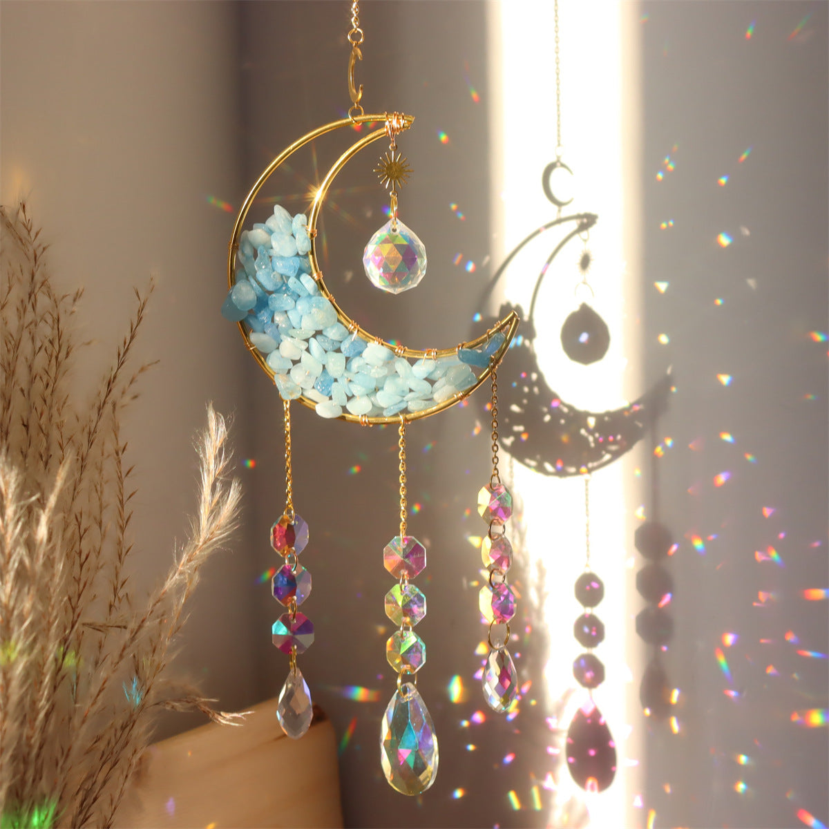 Capteur de Soleil Amour Lunaire en forme de croissant de lune dorée rempli de petites pierres bleues, orné de cristaux arc-en-ciel et d'une boule de cristal prismatique.