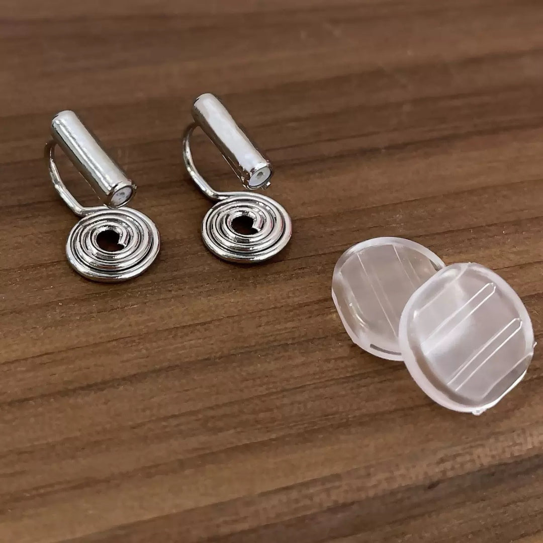 Deux clips d'oreilles argentés en forme de spirale élégante, accompagnés de deux coussinets transparents pour le confort, posés sur un fond en bois sombre.