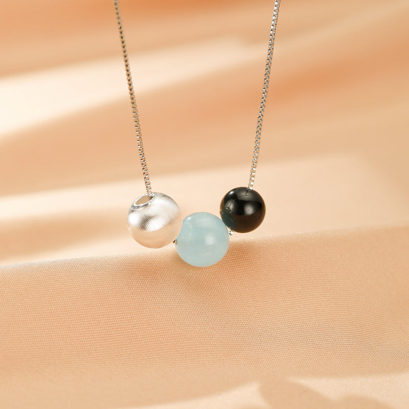Collier "Onde Lumineuse" en argent 925, orné de trois perles : une perle texturée argentée, une perle d'aigue-marine bleu clair et une perle d'obsidienne noire.
