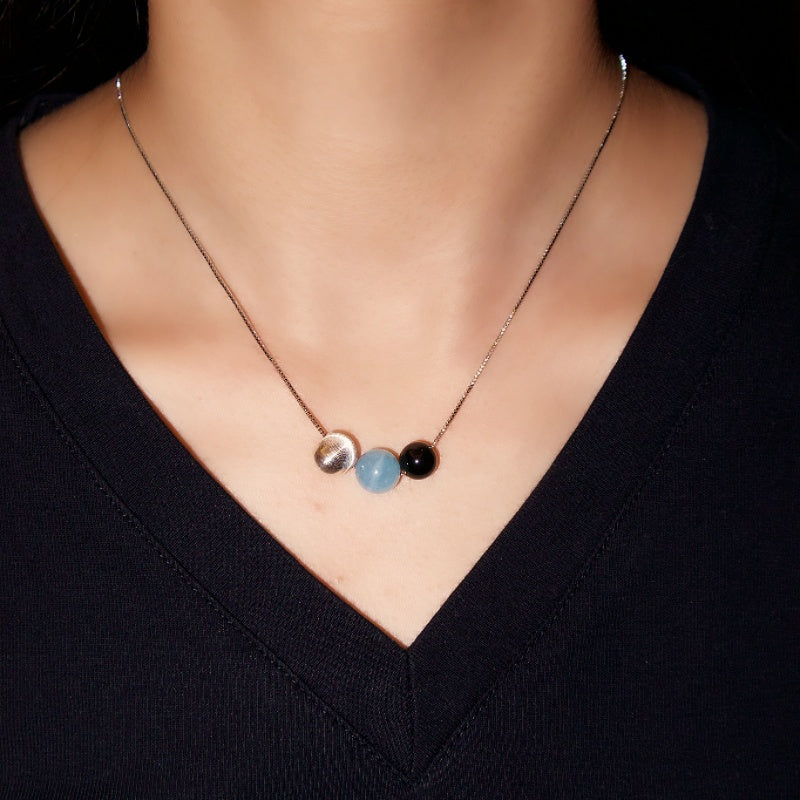 Collier "Onde Lumineuse" en argent 925 orné de trois perles : une perle argentée, une aigue-marine bleu clair et une obsidienne noire, porté par une femme.
