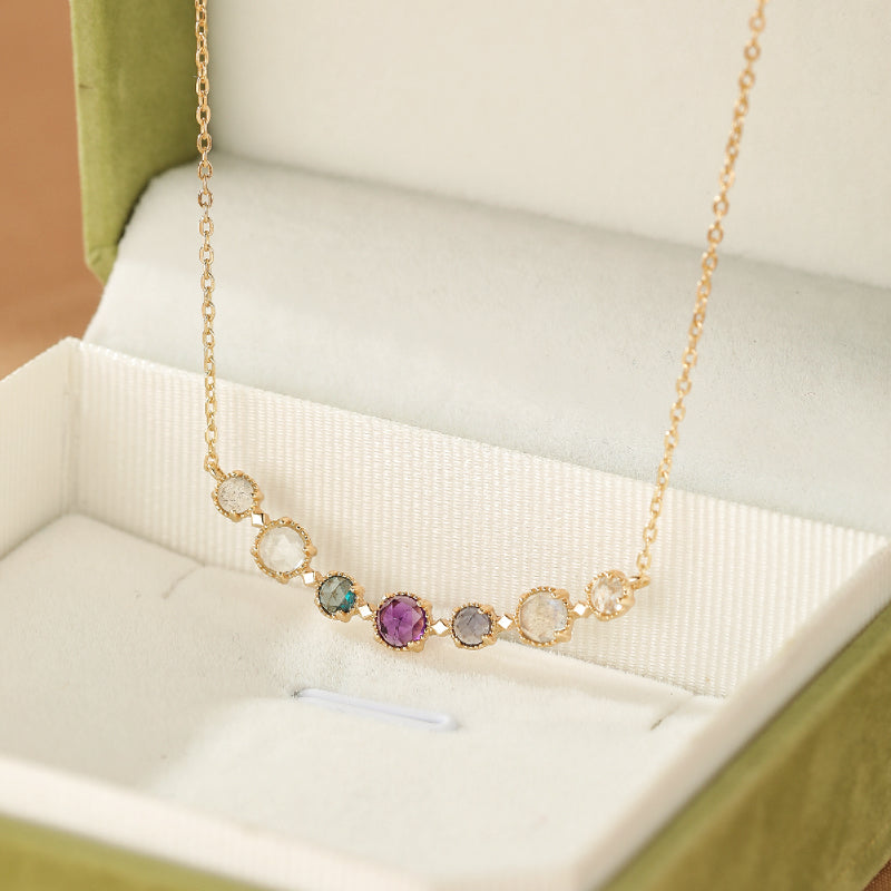 Collier plaqué or "Enchanteur" avec une rangée de pierres naturelles multicolores, dont une améthyste, disposées en arc sur une chaîne fine, présenté dans un écrin vert.
