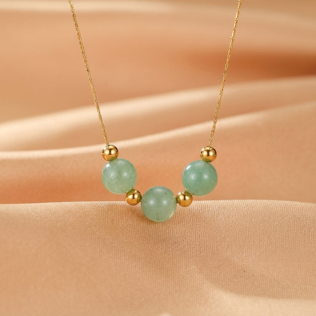 Collier plaqué or avec trois perles rondes d'aventurine verte pâle, espacées par de petites perles dorées, sur un fond en tissu satiné beige.
