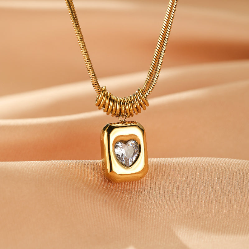Collier doré avec un pendentif rectangulaire et un cœur en cristal au centre, sur une chaîne serpent texturée.