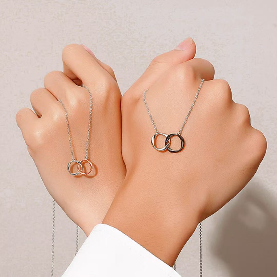 Deux colliers assortis pour couple, l'un en argent et l'autre en argent noirci, chacun avec deux anneaux entrelacés symbolisant l'union.