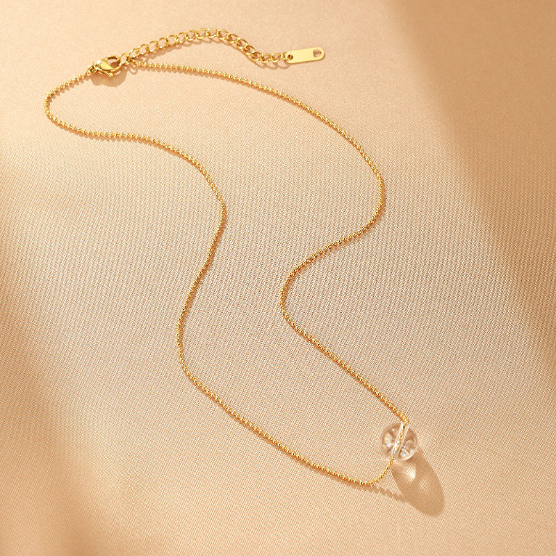 Collier minimaliste en or avec une bille de cristal de roche transparente, monté sur une chaîne à billes dorée, posé sur un tissu beige satiné.