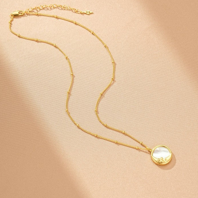 Collier en or jaune avec un pendentif rond en nacre blanche sur fond rose pâle.