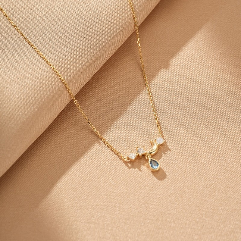 Collier avec un pendentif en forme de lune et de goutte d'eau bleu, orné de petites pierres scintillantes, chaîne fine en argent plaqué or 14k sur fond beige.