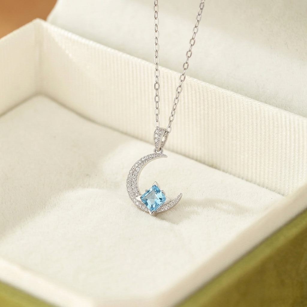 Collier en argent avec un pendentif en forme de croissant de lune orné de petites pierres scintillantes et d'une topaze bleue carrée au centre.