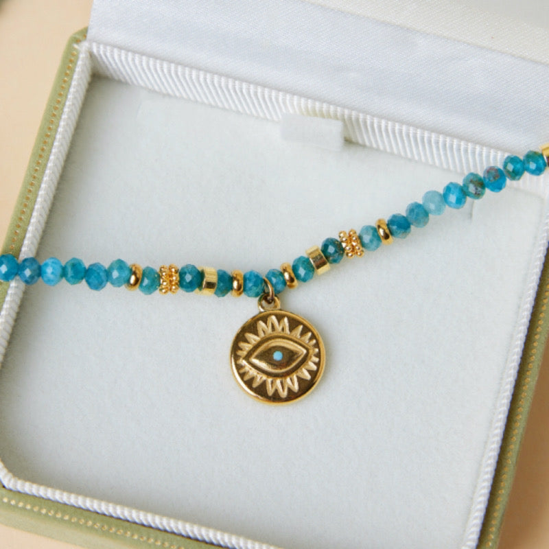 Gros plan sur un collier bleu avec des perles d'apatite et un pendentif œil doré, posé sur un coffret cadeau.