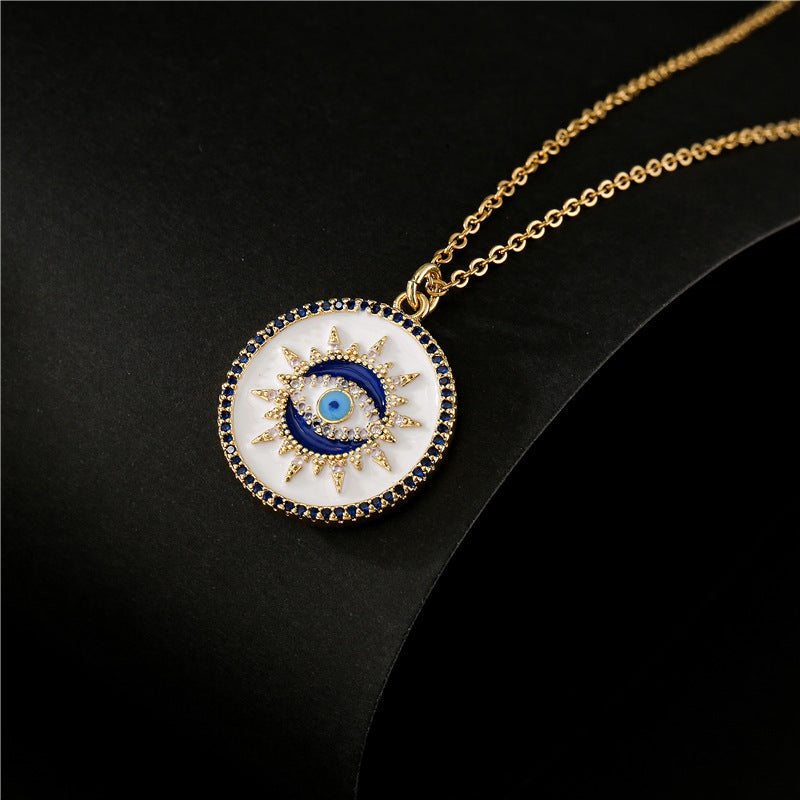 Collier doré avec pendentif rond blanc orné d'un œil bleu stylisé, d'un croissant de lune bleu et de cristaux blancs et bleus, symbolisant la protection.